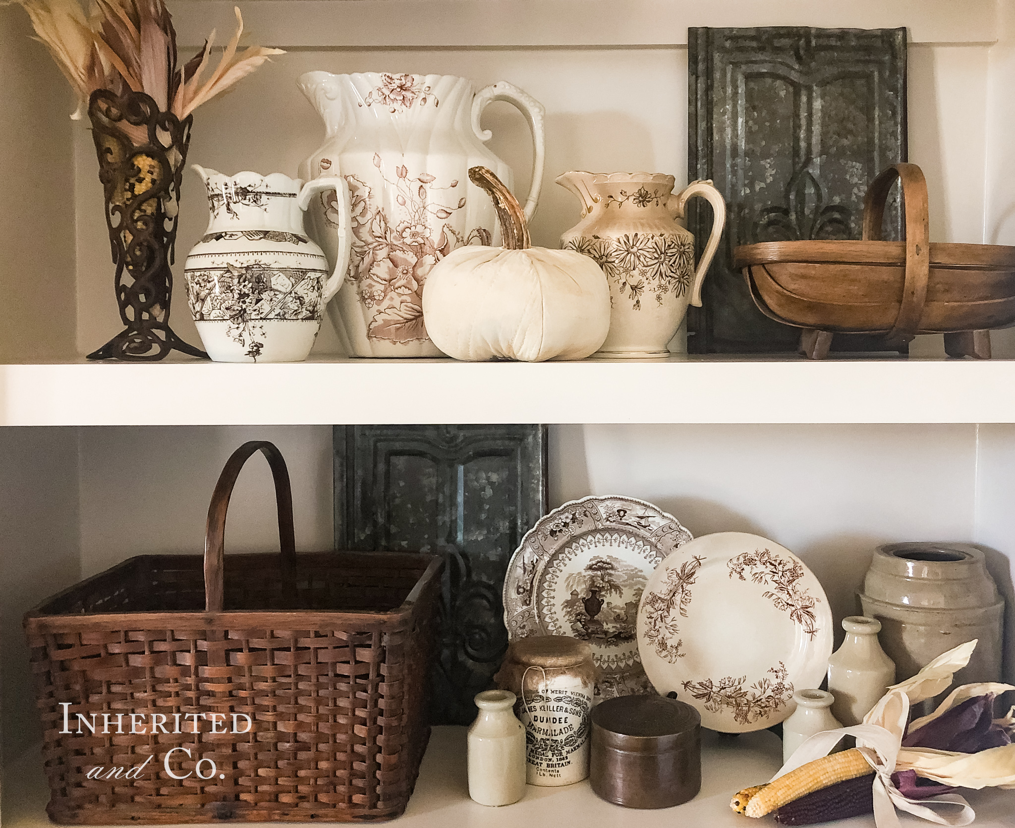 Two built-in white shelves filled with antiques such as brown transferware baskets stoneware weathered metal copper dried corn and a quilt pumpkin