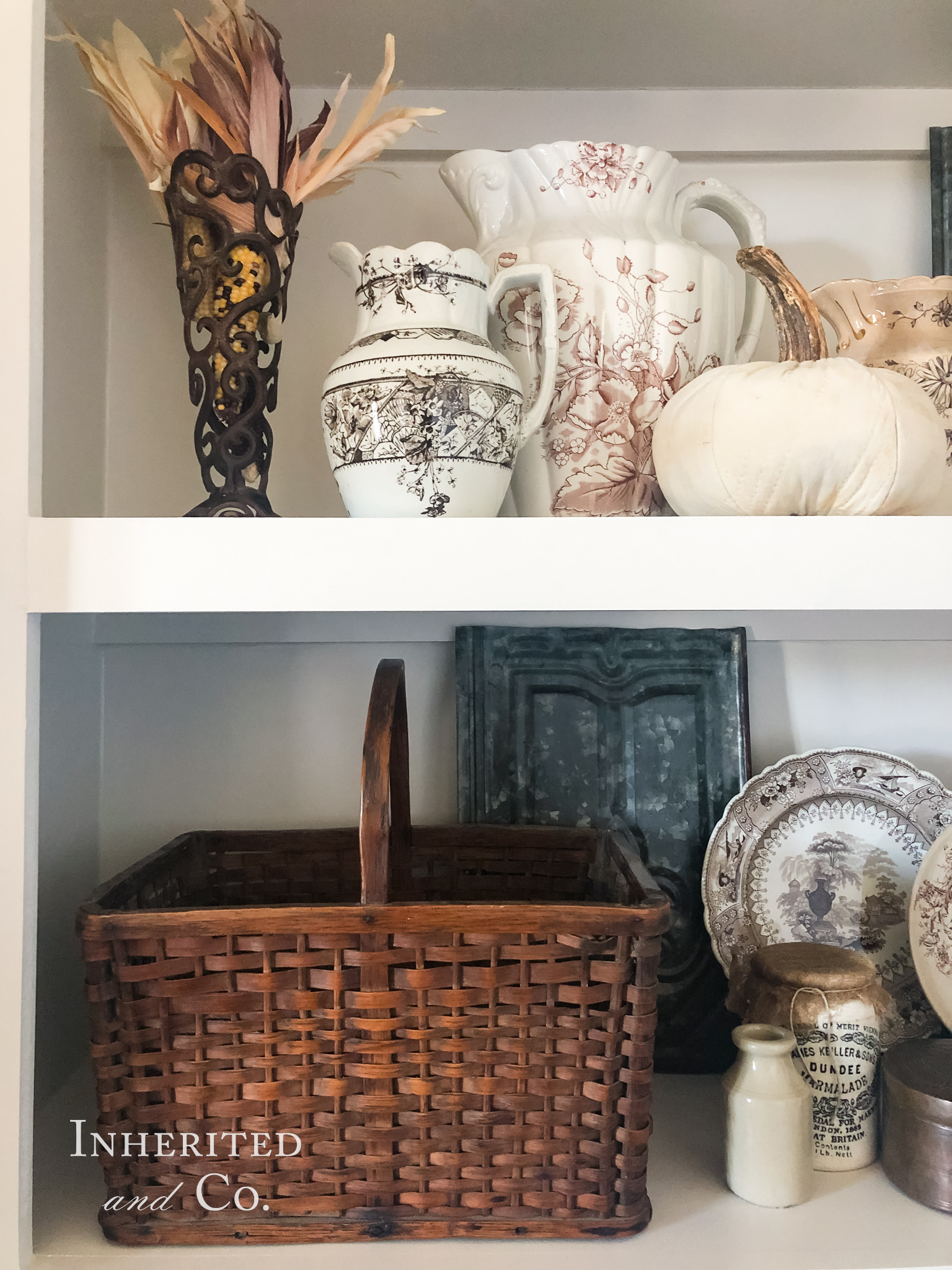 Two white built-in shelves filled with antiques such as brown transferware plates and pitchers, stoneware, harvest baskets, and weathered metal