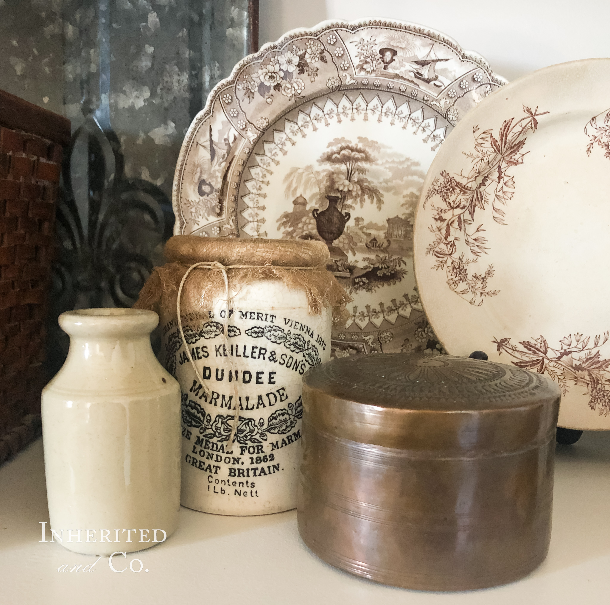 An antique stoneware ink bottle, Dundee marmalade jar, and round copper lidded box in the foreground with two antique brown transferware plates in the background