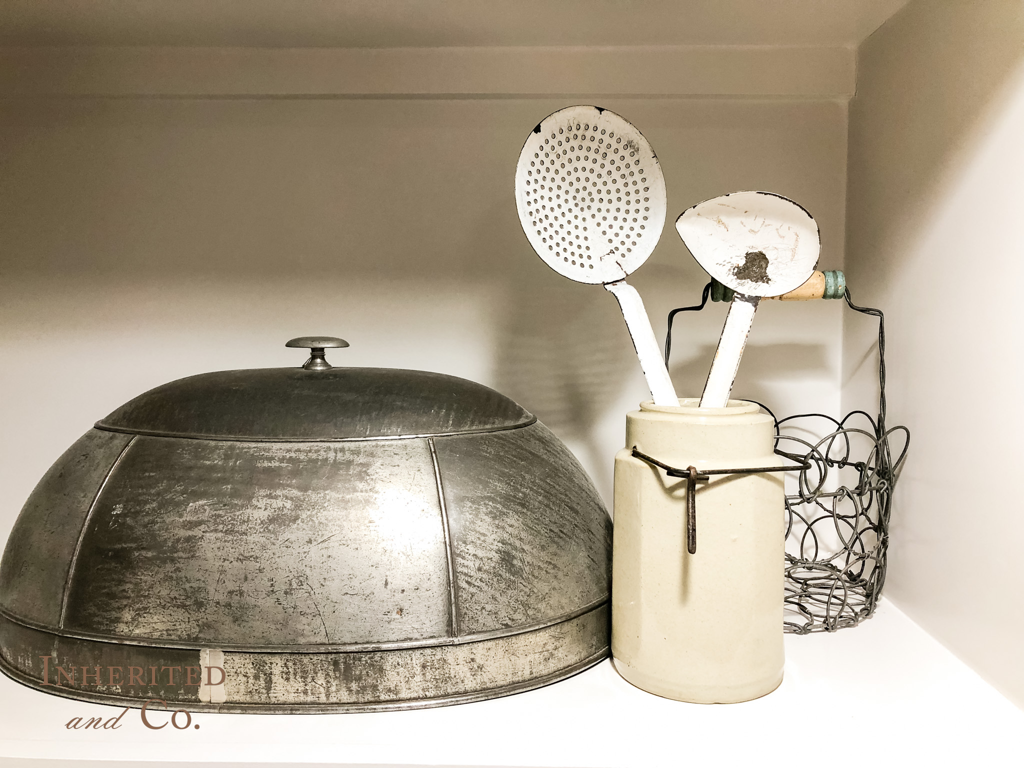 vignette of antiques , including meat dome, stoneware crock, enamel utensils, and wire egg basket