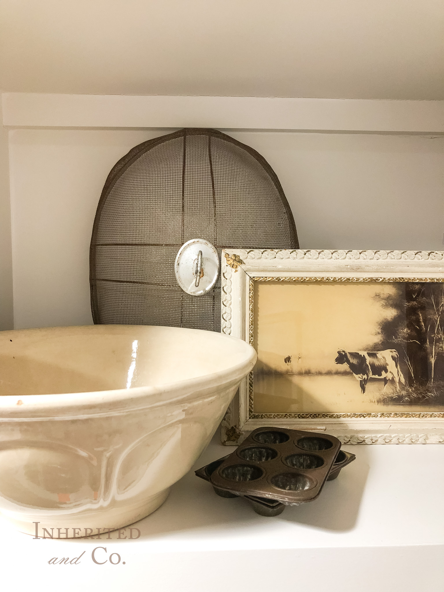 vignette of antiques including an ironstone bowl, baking pans, meat dome, and framed cow print